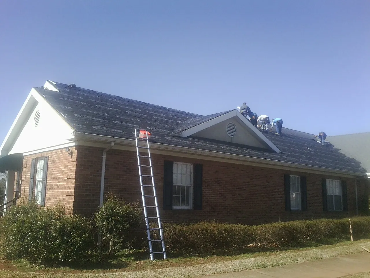 Crew tearing shingles off a brick ranch roof with a ladder set — Emergency Roof Repair work by our crew in Brent