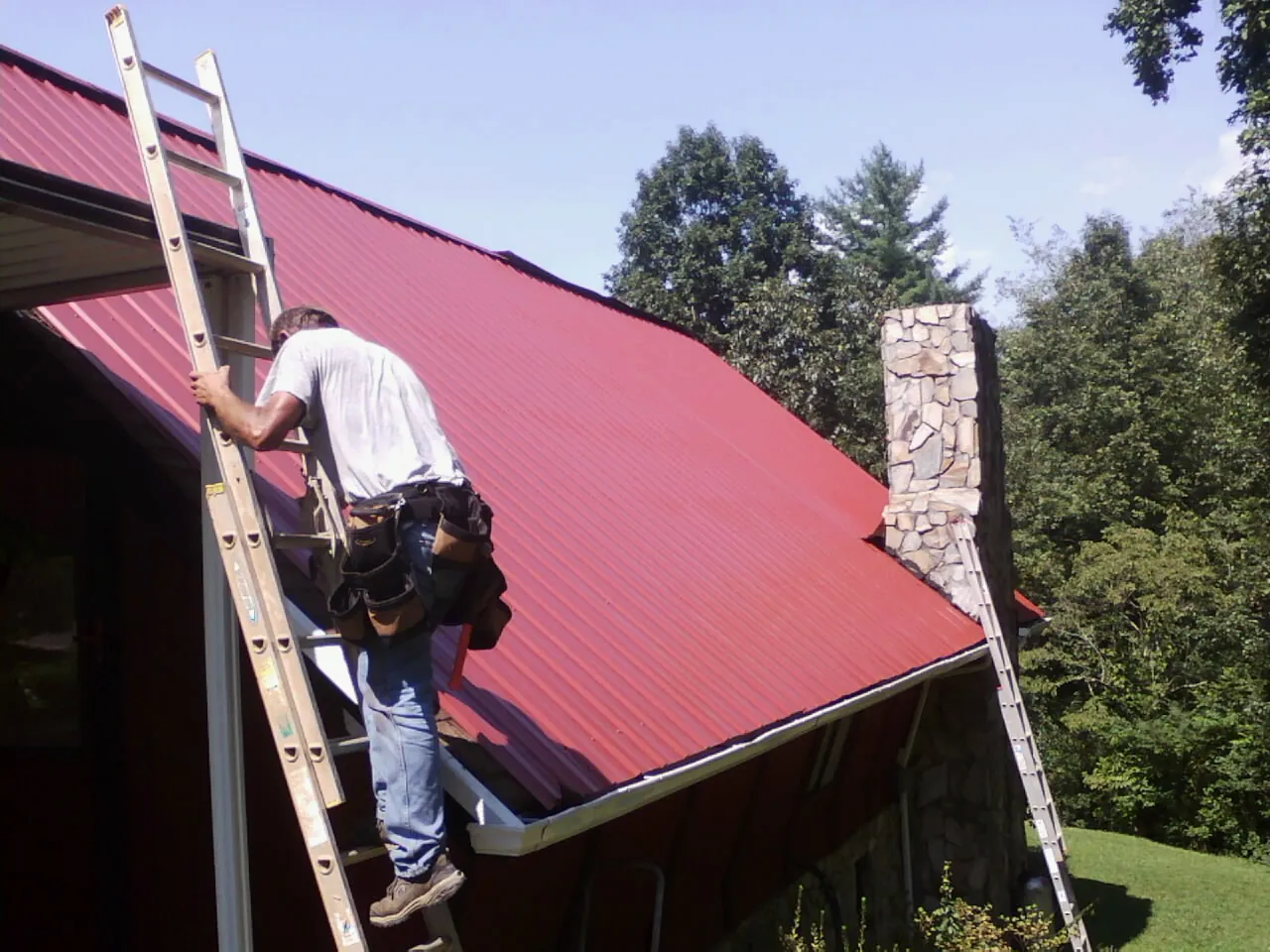 Roofer on a ladder beside a red metal roof and stone chimney — Soffit & Fascia Repair by Roofing Brothers in Brent